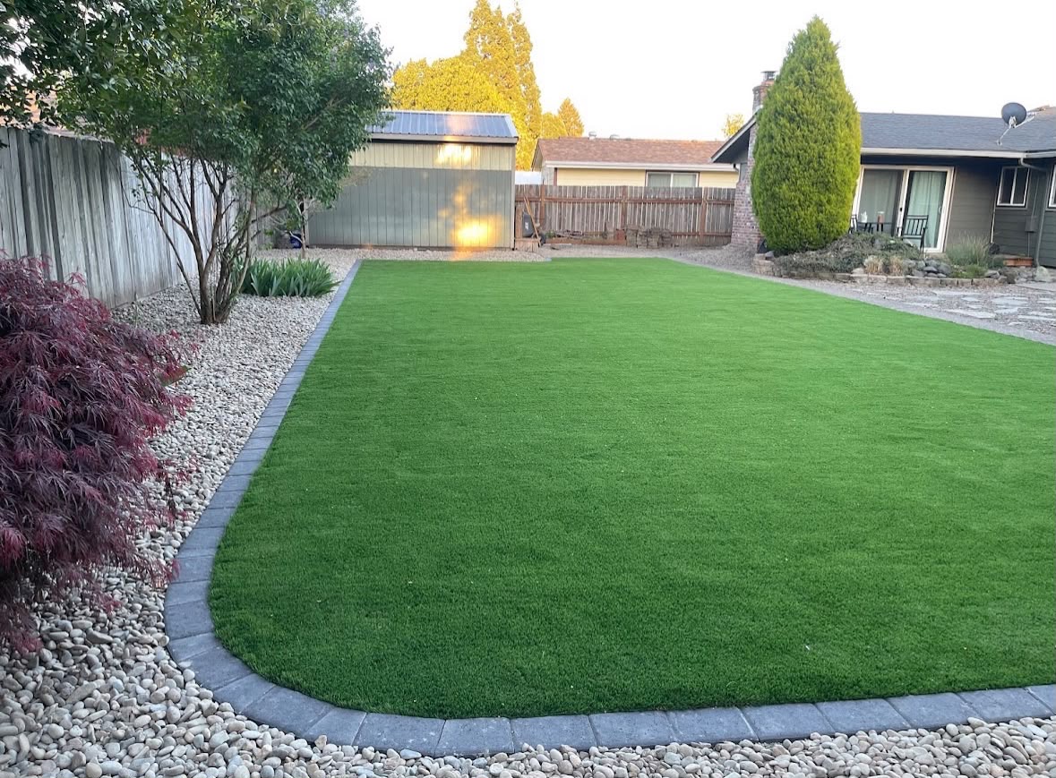 Artificial turf lawn with a charcoal paver border and river rock surround — GI Landscape