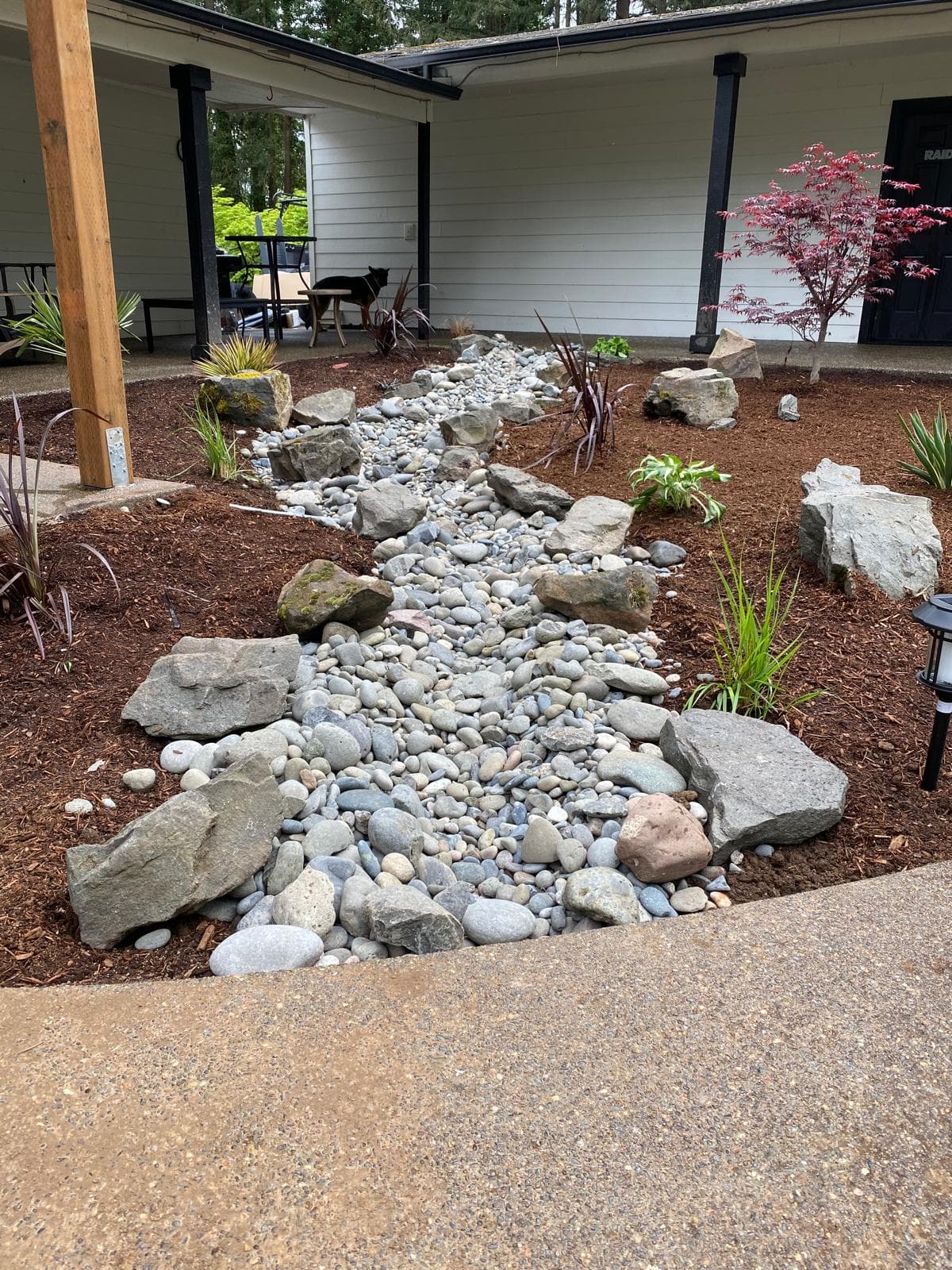 Dry creek bed — a decorative river-rock drainage swale with boulders beside a covered patio, by GI Landscape