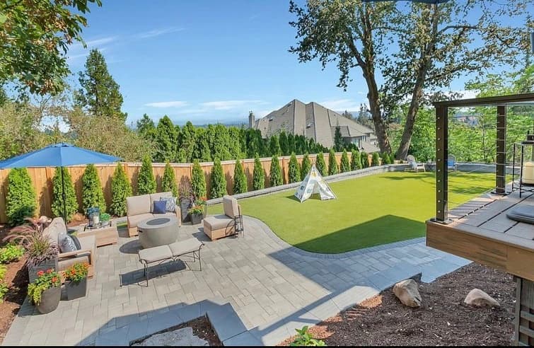 Aerial view of a finished GI Landscape backyard — paver patio with outdoor seating, artificial turf lawn, cable-rail glass deck, with neighborhood and mountain horizon in the distance